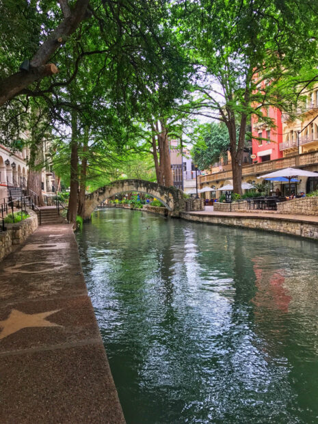 A serene view of San Antonio riverwalk with lush green trees and stone bridge