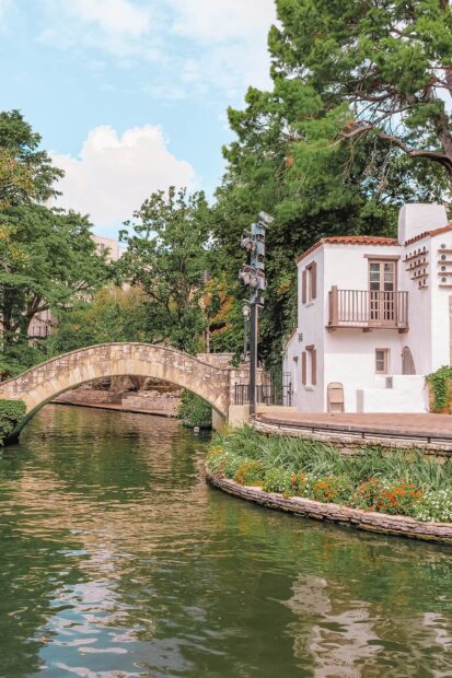 A charming stone bridge over a river in San Antonio Texas with greenery and a white building nearby