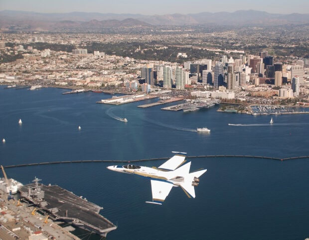 A jet flying over the San Diego skyline and harbor with mountains in the background