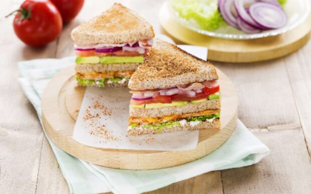 A fresh sandwich with layers of tomato avocado onion and lettuce on toasted bread placed on a wooden board