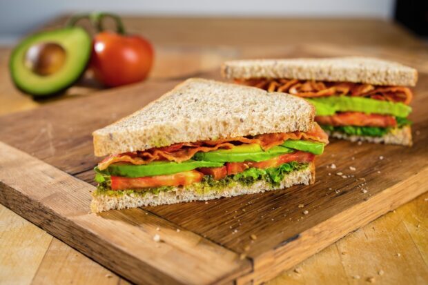 A fresh sandwich with avocado bacon and tomato on whole grain bread on a wooden cutting board
