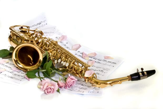 A golden saxophone laying on music sheets surrounded by pink roses and petals