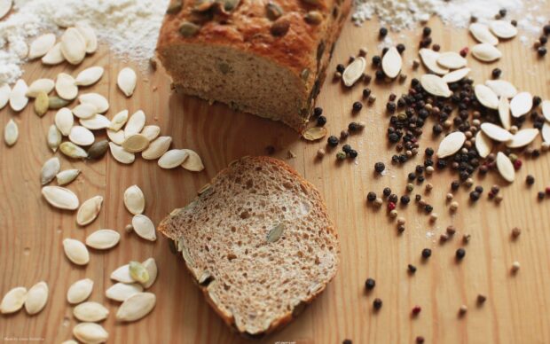 A slice of bread with seeds surrounded by scattered seeds on a wooden surface