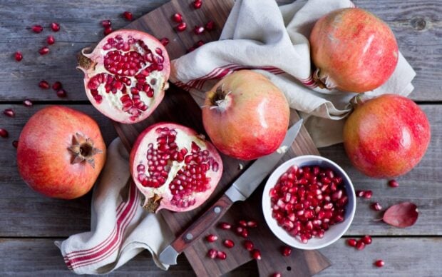 Fresh pomegranate seeds and whole fruits arranged on a wooden surface with a knife and cloth