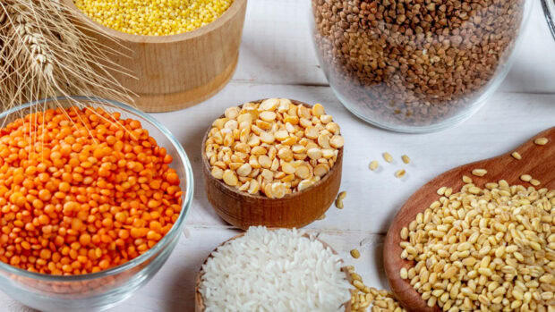 Various seeds in bowls and jars on white wooden surface with wheat stalks and rice grains