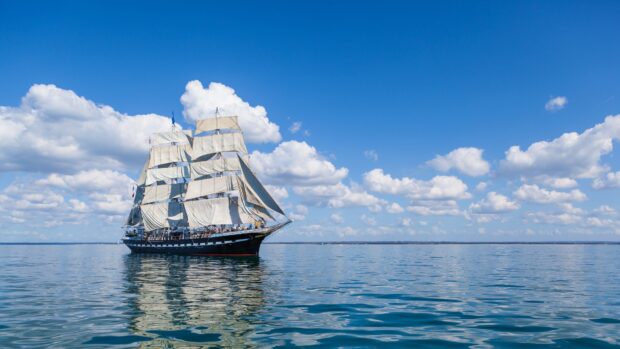 A classic tall ship sailing on calm ocean water under a blue sky with white clouds