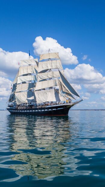 A tall sailing ship with white sails reflecting on calm water under a blue sky with clouds