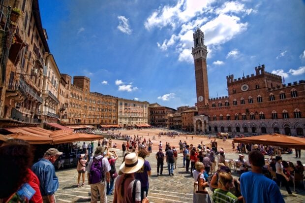 Crowded square with historic buildings and tower in Siena city center on a clear sunny day