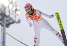 A ski jumper in midair wearing a red helmet and white suit during a competition
