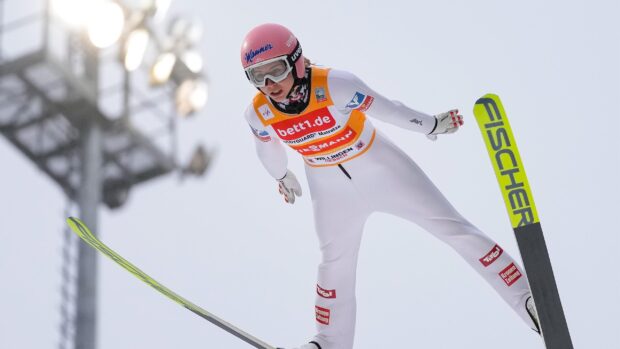A ski jumper in midair wearing a red helmet and white suit during a competition