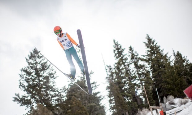 A ski jumper in mid air wearing an orange helmet and green pants above snow and trees