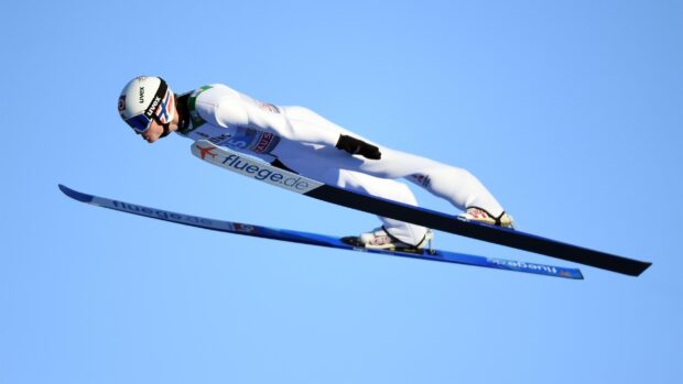 A ski jumper wearing a white suit soaring through the air against a clear blue sky