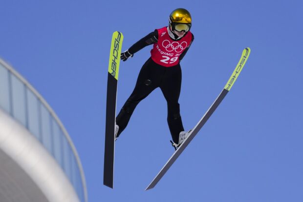 Ski jumping athlete wearing a golden helmet soaring through the air against a clear blue sky