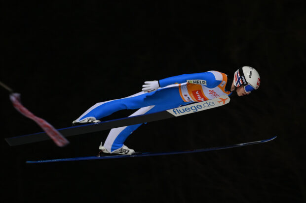 A ski jumper in blue and white suit soaring through the air during a night ski jumping event