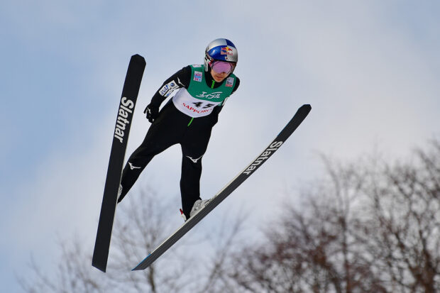 Male ski jumper in flight wearing helmet and goggles over winter trees