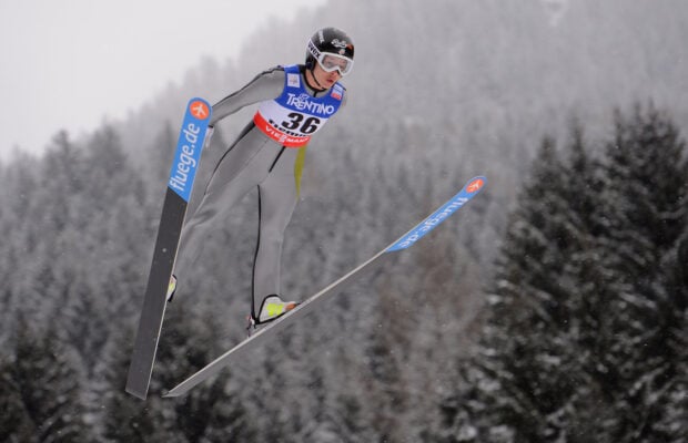 Ski jumping athlete in gray suit soaring above snow covered forest trees