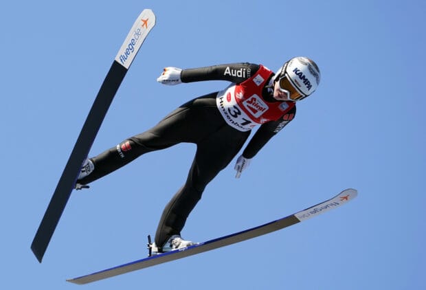 A ski jumper in full flight wearing a black suit and helmet against a clear blue sky