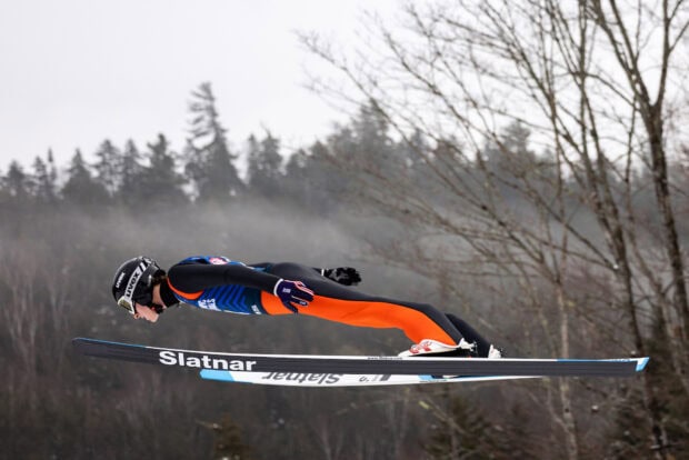 A ski jumper in full flight wearing an orange and black suit over a snowy forest background