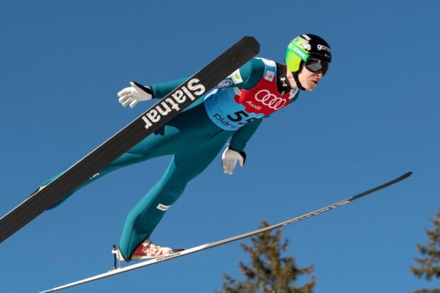 A ski jumper in midair wearing green suit and helmet during a competition against a clear blue sky