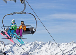 Two skiers wearing colorful gear riding a ski lift with snowy mountain peaks in the background