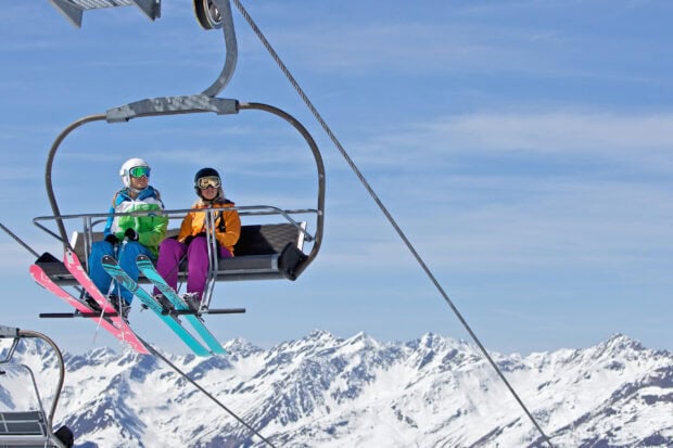 Two skiers wearing colorful gear riding a ski lift with snowy mountain peaks in the background