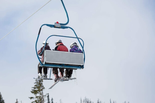 Three skiers wearing helmets on a ski lift surrounded by snow and trees