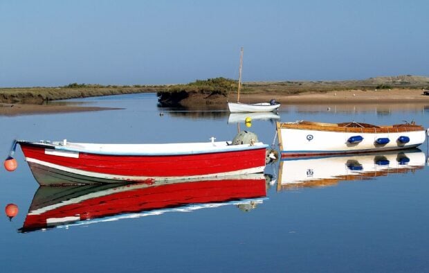 A serene view of a red skiff floating calmly on clear water with reflections and surrounding marshland