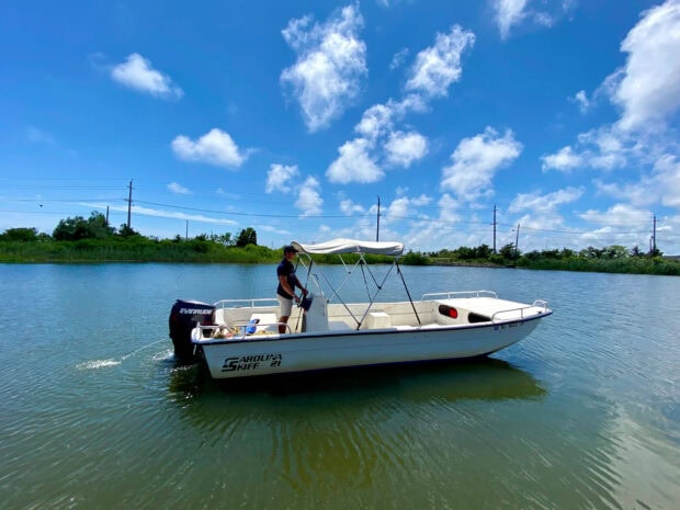 A man steering a skiff boat on calm water under a bright blue sky with scattered clouds