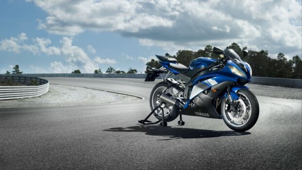 A blue sports bike parked on a race track under a cloudy sky with trees in the background