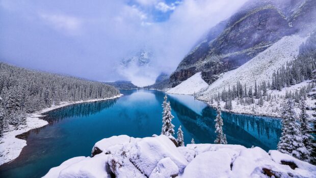 Snow covered spruce trees surround a clear blue lake in a winter mountain landscape