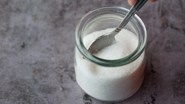 A jar filled with white sugar granules and a metal spoon inside on a gray surface