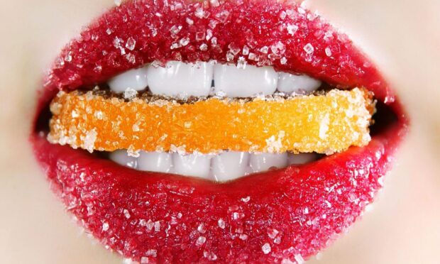 Red lips with sugar crystals holding an orange candy piece between white teeth