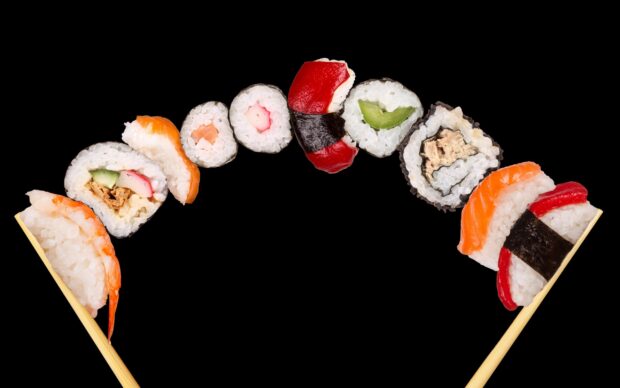 An assortment of fresh sushi rolls arranged in an arc held by chopsticks on a black background