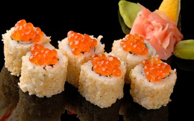 Sushi rolls topped with salmon roe and sesame seeds served with ginger and wasabi on black surface