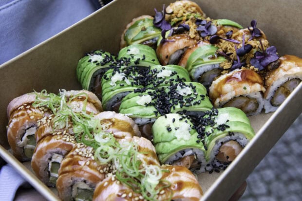 Fresh sushi roll topped with avocado and black sesame seeds in a takeout box