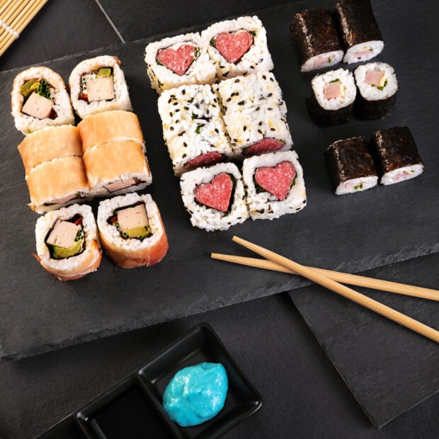 Various sushi rolls with heart shaped meat filling served on a black slate plate with chopsticks and blue sauce