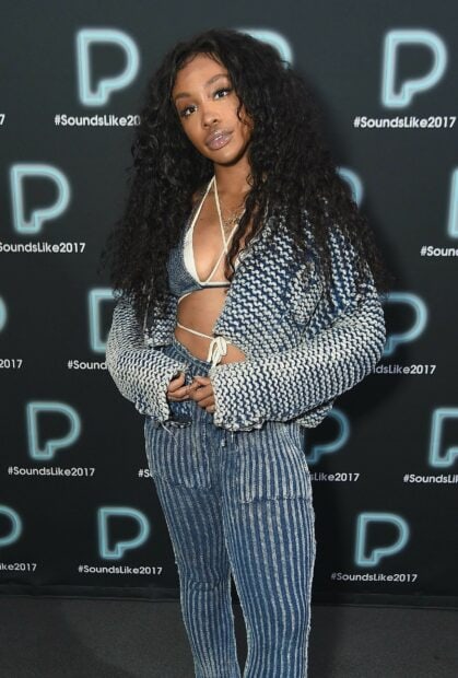 Sza wearing a knitted jacket and striped pants posing at an event with a branded backdrop