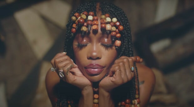 Sza with braided hair and wooden beads resting her face on her hands with eyes closed