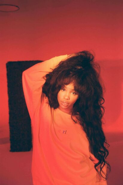 A portrait of Sza wearing a casual long sleeve shirt with voluminous hair in a warm red setting