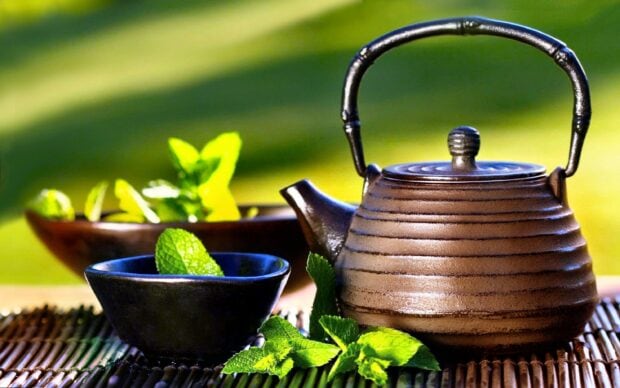 A brown teapot with fresh mint leaves in a bowl and on a bamboo mat
