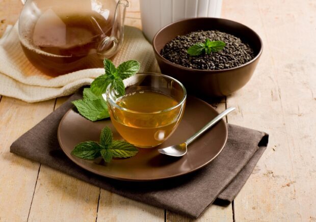 A glass cup of green tea with fresh mint leaves and a bowl of tea leaves on a wooden table