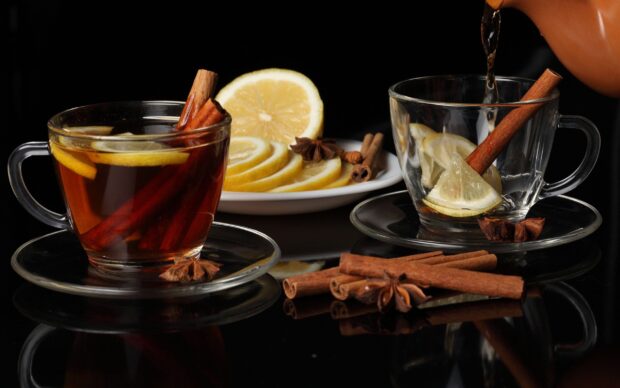 A glass cup of tea with cinnamon sticks and lemon slices on a reflective black surface