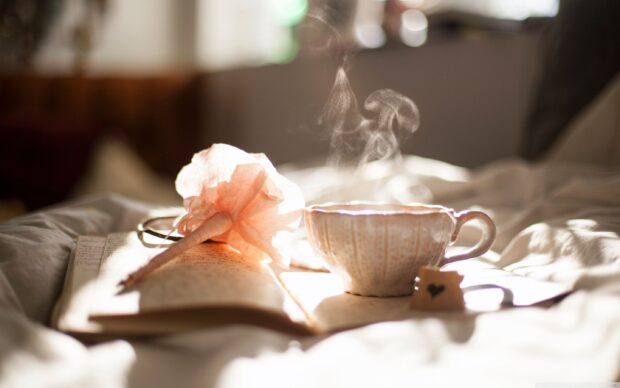 A steaming cup with tea tag beside a book and a decorative pen on a cozy bed