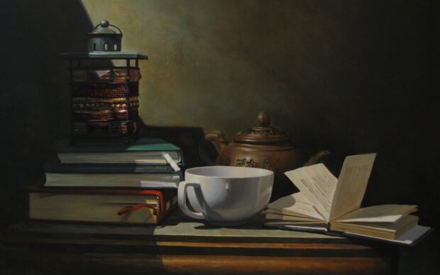 A tea cup next to a teapot with books and a lantern on a wooden table in soft lighting