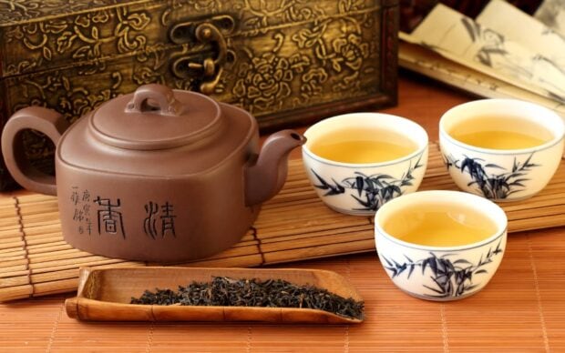 A traditional teapot and three cups of brewed tea with loose tea leaves on a bamboo mat