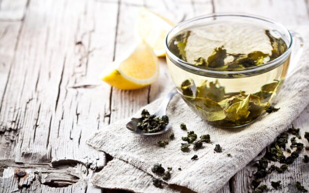 A cup of green tea with loose tea leaves and lemon wedges on a wooden table