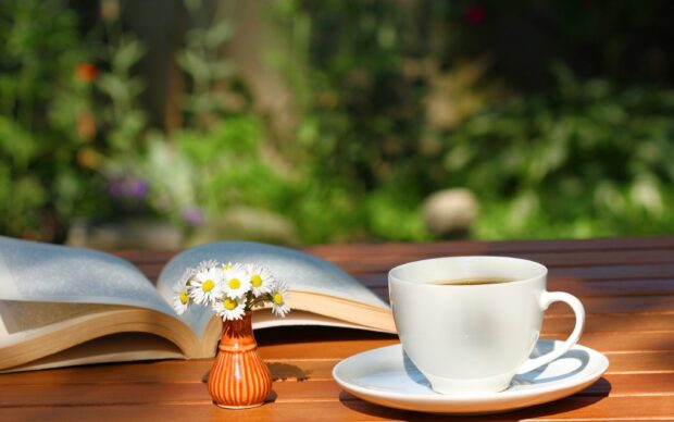 A cup of tea next to a small vase with daisies and an open book on a wooden table