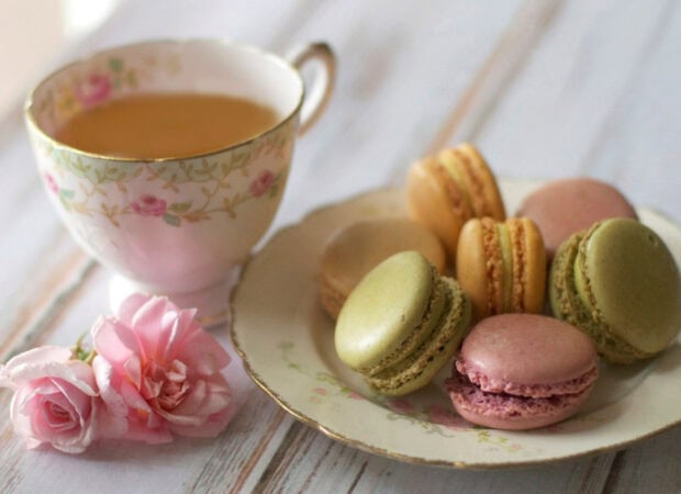 A tea cup with floral design and a plate of colorful macarons with pink roses on a wooden table