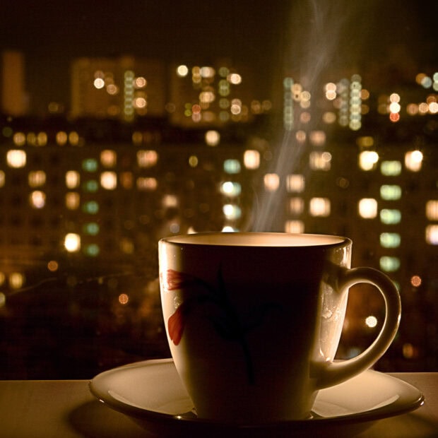 A steaming cup with flower decoration on a saucer in front of city lights at night