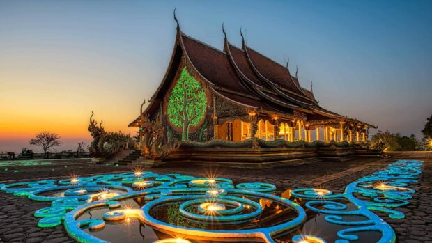 Thai temple with glowing green tree design and blue patterned lights at sunset in Thailand